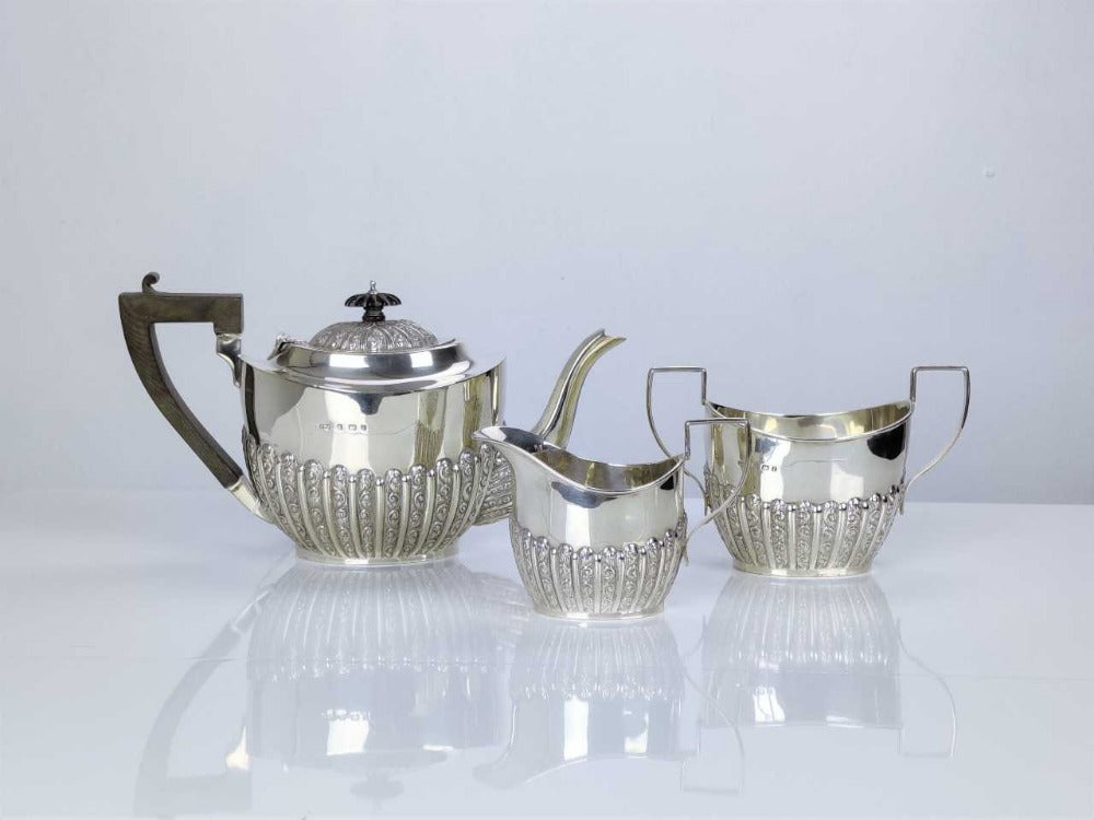 A silver three-piece tea service set including a teapot, a pot for sugar or cream, and a cup, all with intricate designs and a polished finish.