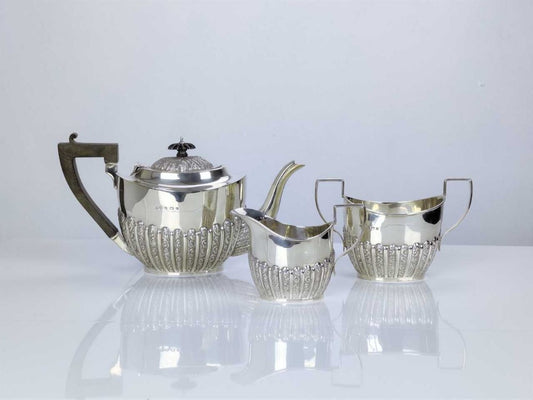 A silver three-piece tea service set including a teapot, a pot for sugar or cream, and a cup, all with intricate designs and a polished finish.